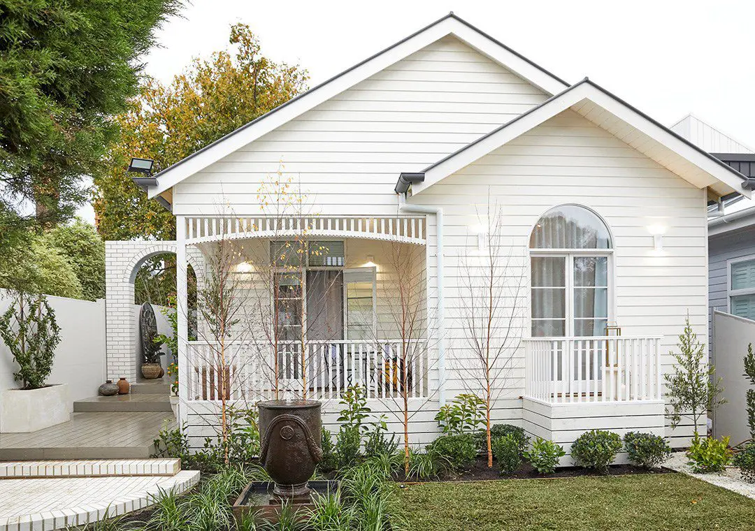 A charming white cottage with James Hardie Fibre Cement siding features a front porch, arched windows, and picket railings. It’s surrounded by a neat garden with shrubs, a small fountain, young trees, and a brick pathway leading to the entrance.
