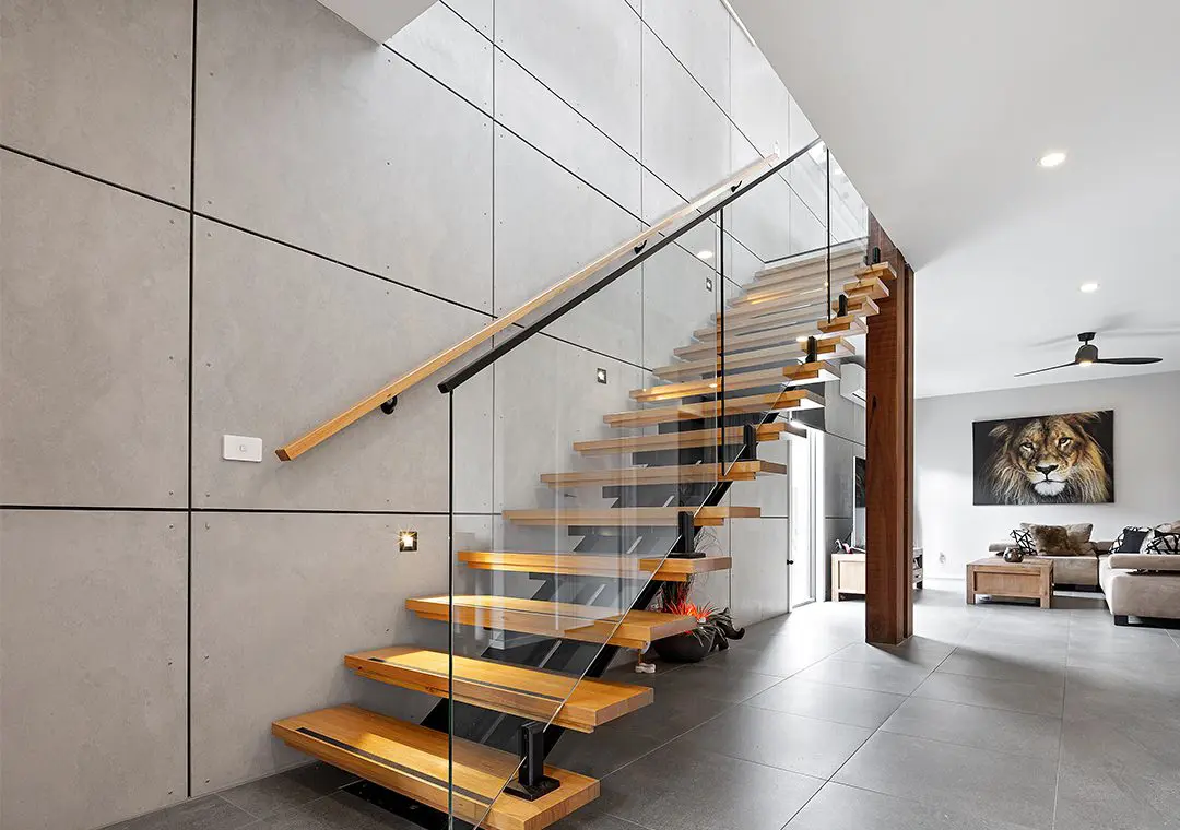 Modern interior with floating wooden stairs and glass railing, grey tiled walls and floor finished with James Hardie Fibre Cement, and a living area in the background featuring a large lion painting, sofa, and ceiling fan. Bright, natural lighting.