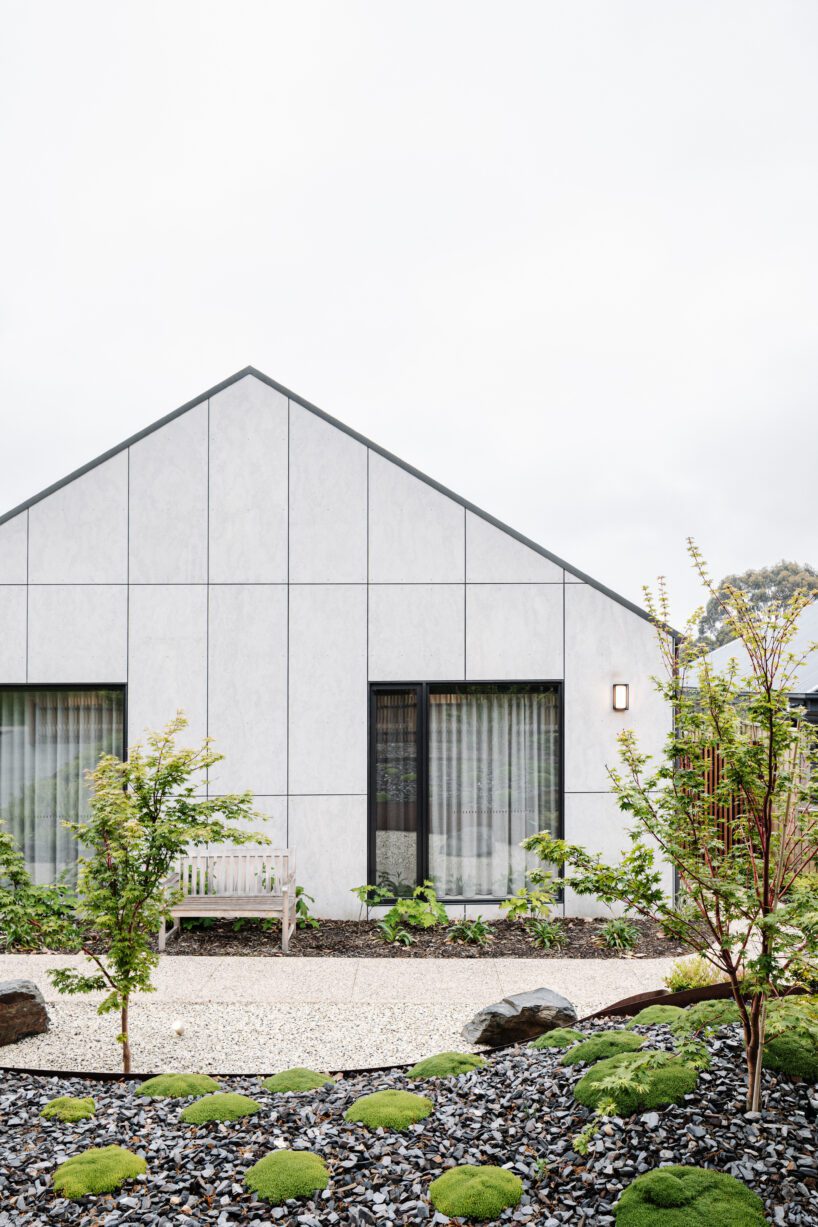 A modern white house with a peaked roof, large windows, and a light fixture features sleek James Hardie Fibre Cement siding. Out front, find a white bench by the gravel path and a landscaped garden with small plants, moss, rocks, and young trees.