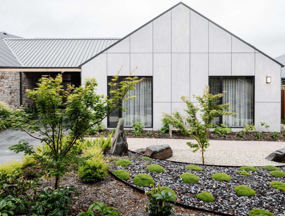 Modern house with a gray, gabled roof and large front windows, featuring James Hardie Fibre Cement exterior, surrounded by a minimalist garden with small trees, green shrubs, rocks, gravel, and neatly arranged landscaping.