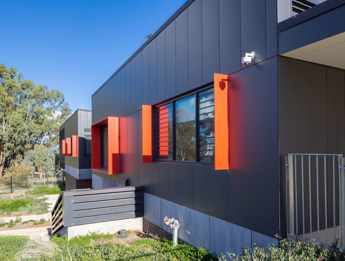Modern building exterior with dark panels, bright orange window frames, and large windows featuring horizontal slats; clad in James Hardie Fibre Cement, with a security camera and landscaped greenery beneath a clear blue sky.