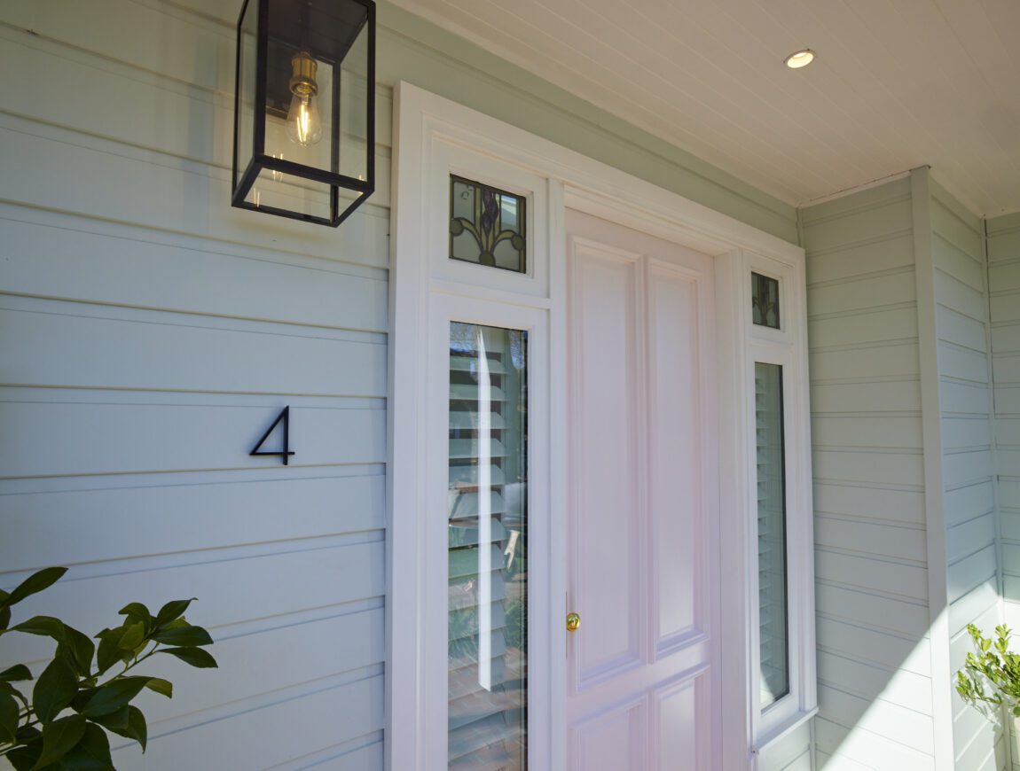 A pale blue James Hardie Fibre Cement house exterior with white trim, featuring a white front door flanked by narrow windows. A black lantern-style light fixture hangs above the number 4 next to the door. Green plants are partially visible.