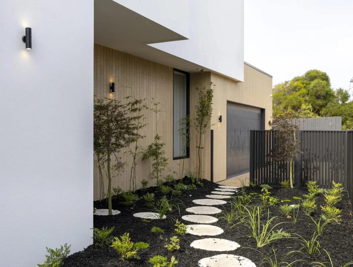 Modern house exterior with white and light wood James Hardie Fibre Cement walls, circular stepping stones in a black-mulched garden, young plants, minimalist fence, and garage door visible.