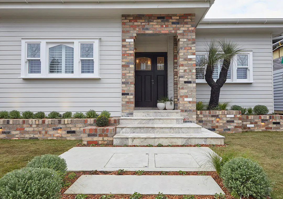 Single-story house entrance with James Hardie Fibre Cement gray siding, brick porch, black front door, small windows with shutters, two potted plants by the door, neatly trimmed bushes, and a tree in a landscaped yard.