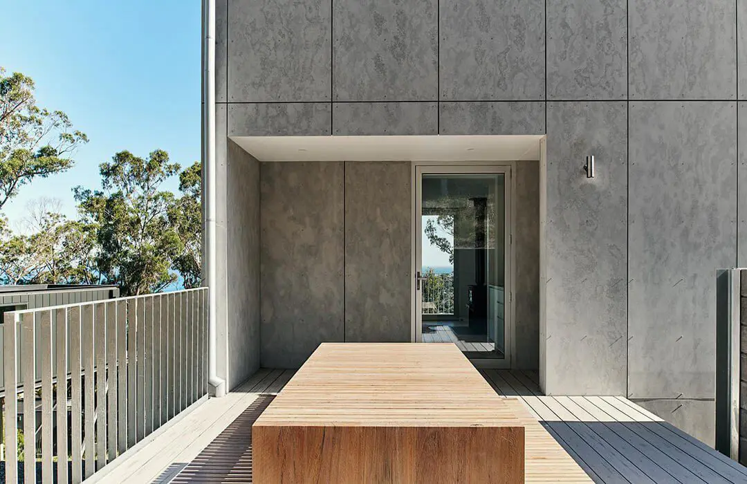 A modern patio with James Hardie Fibre Cement gray concrete walls, a wooden table in the center, metal railings, and trees visible in the background under a clear blue sky.