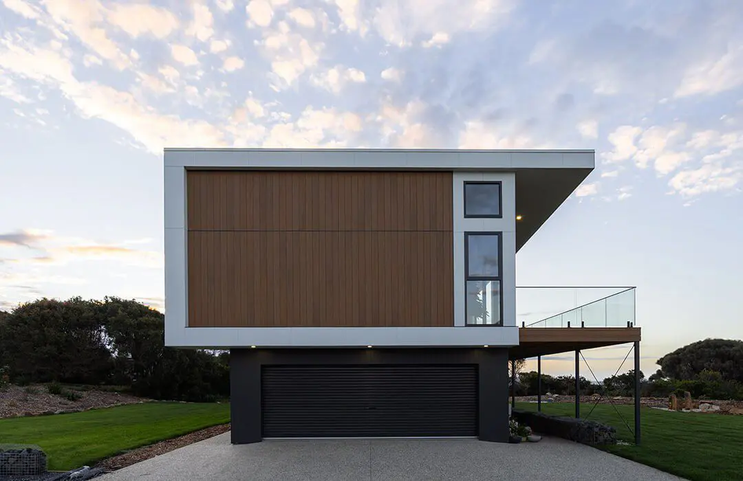 A modern, box-shaped house with James Hardie Fibre Cement wood paneling, large windows, and an elevated structure supported by columns sits above a dark garage. The sky is partly cloudy, and landscaping surrounds the property.