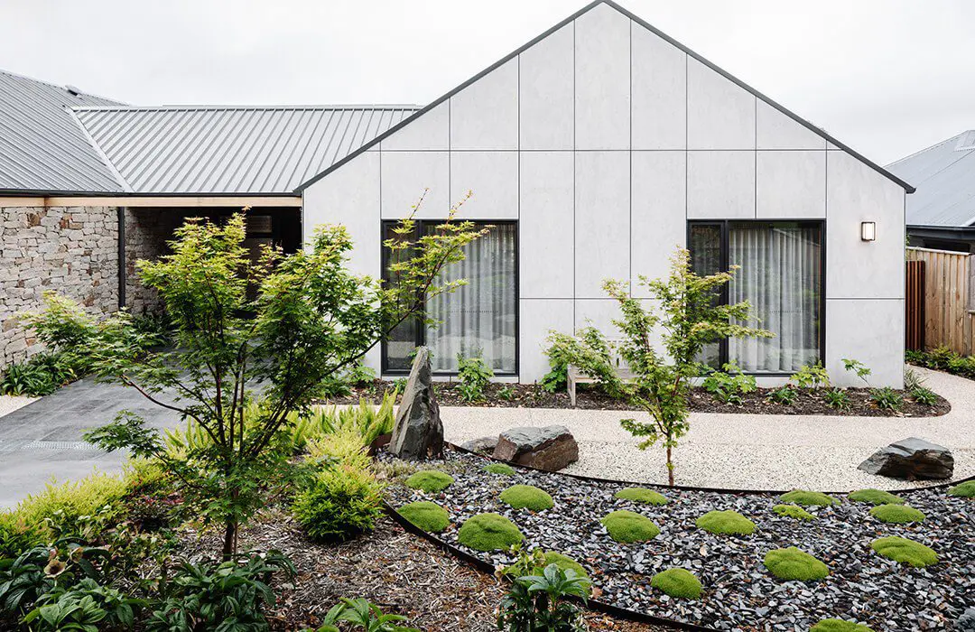 Modern house with light gray James Hardie Fibre Cement panels, two large windows, and a minimalist front yard featuring small trees, green shrubs, rocks, and a gravel pathway. The clean landscaping enhances the home’s contemporary look.