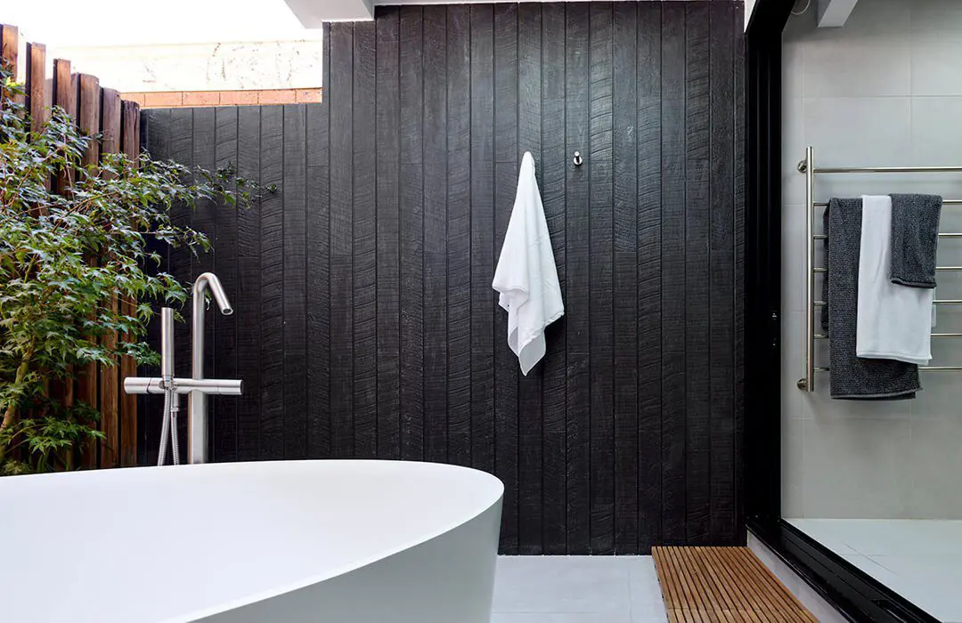 A modern outdoor bathroom with a white freestanding tub, black James Hardie Fibre Cement wood-paneled wall and white towel, a tall plant, and towels on a rack next to a glass shower door.