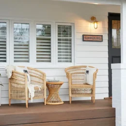 Two wicker chairs with cushions and a small wicker table sit on a wooden porch outside a white house clad in James Hardie Fibre Cement, featuring three windows, a black front door, and a sign reading SUMMERHILL by the entrance.
