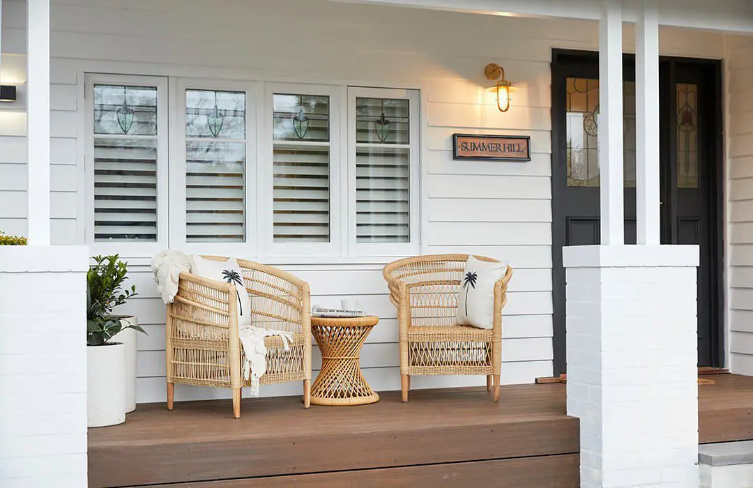 Two wicker chairs with cushions and a small wicker table sit on a wooden porch outside a white house clad in James Hardie Fibre Cement, featuring three windows, a black front door, and a sign reading SUMMERHILL by the entrance.