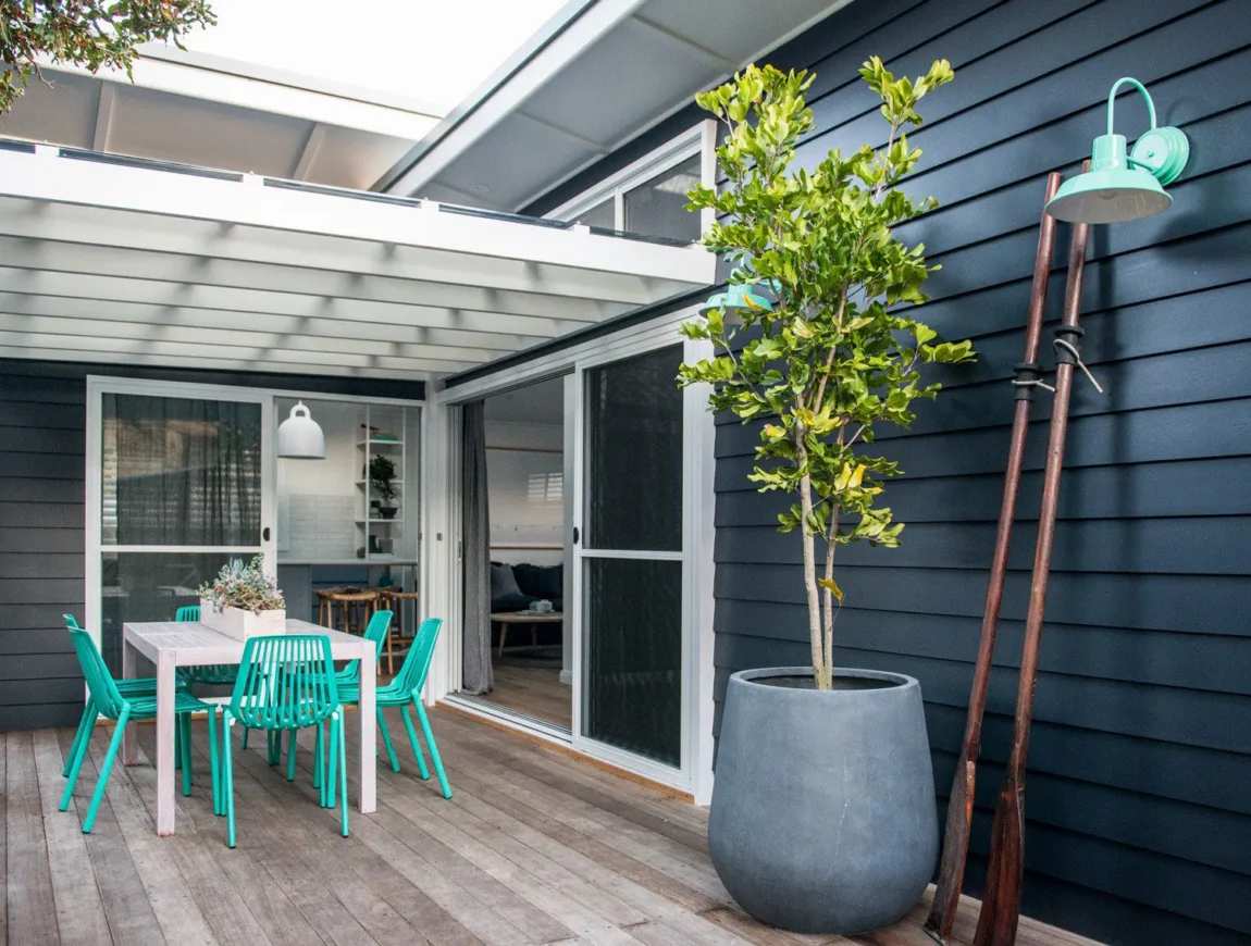 A modern outdoor patio with a wooden deck, turquoise table and chairs, large potted plant, navy James Hardie Fibre Cement horizontal siding, sliding glass doors, and a green wall-mounted light fixture.