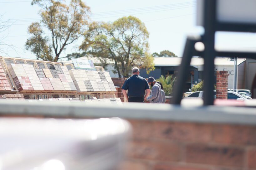 Two people stand outdoors near tile displays, possibly at a hardware store like MD Brick Central Coast. Trees and parked cars are visible in the background on a sunny day, while the foreground is partially obstructed by a blurry structure.