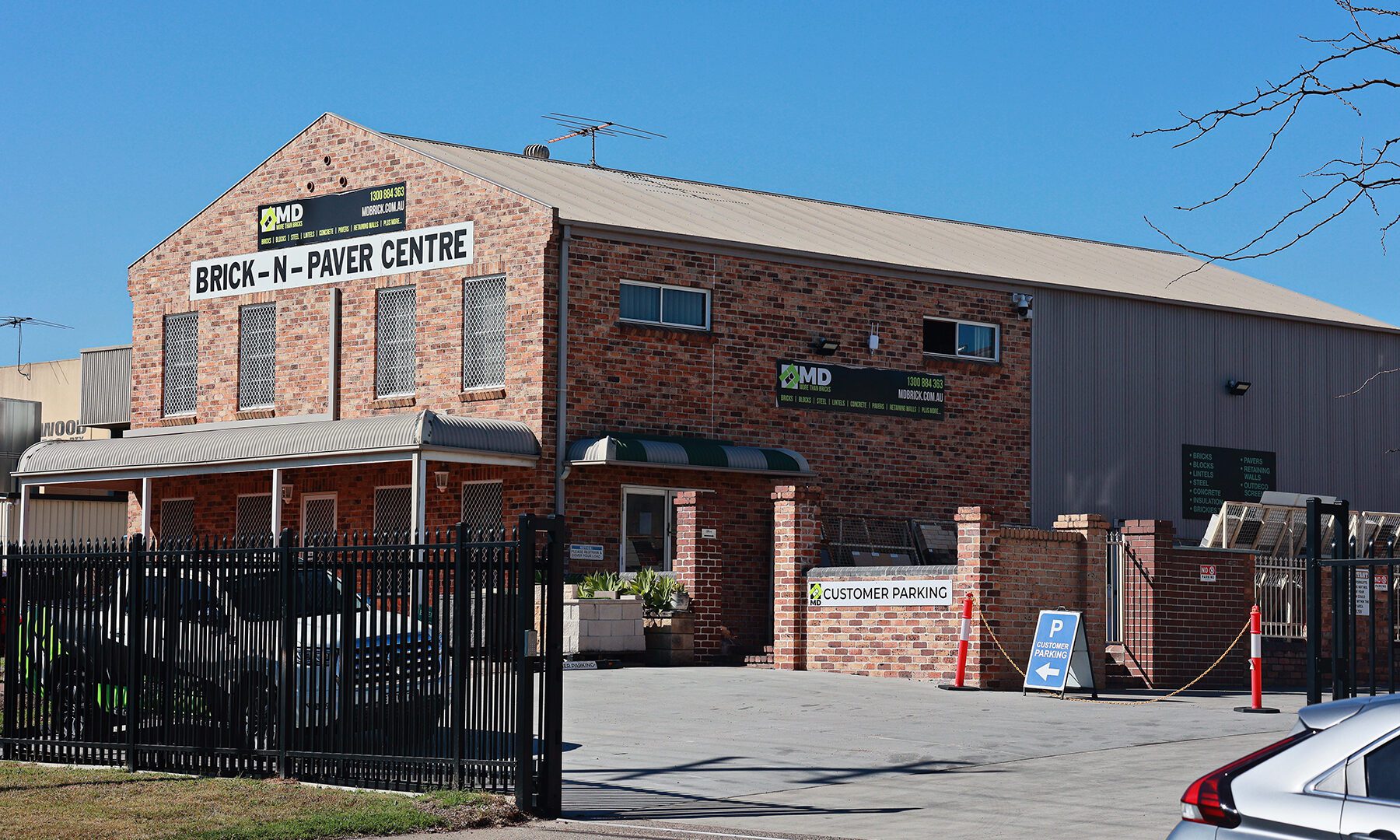 A brick building labeled BRICK-N-PAVER CENTRE, part of MD Brick Central Coast, features a fenced parking lot, signs for customer parking, and a clear blue sky in the background.