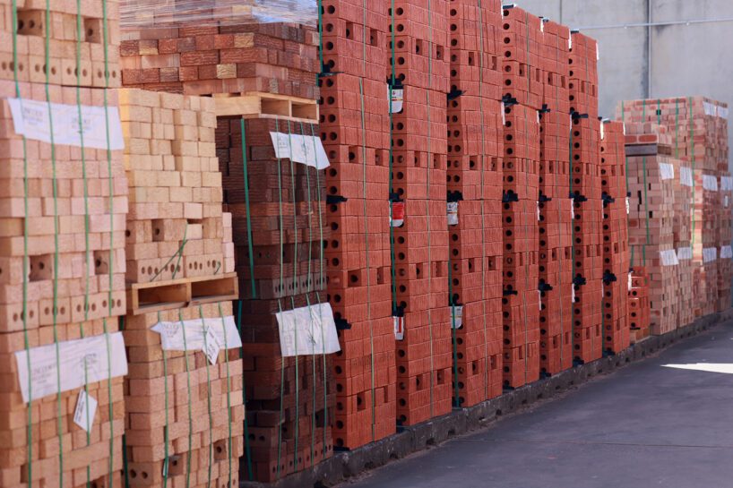 Stacks of red and light brown bricks are neatly arranged on pallets in an outdoor storage area at MD Brick Central Coast, ready for construction use. The bricks are secured with green straps.