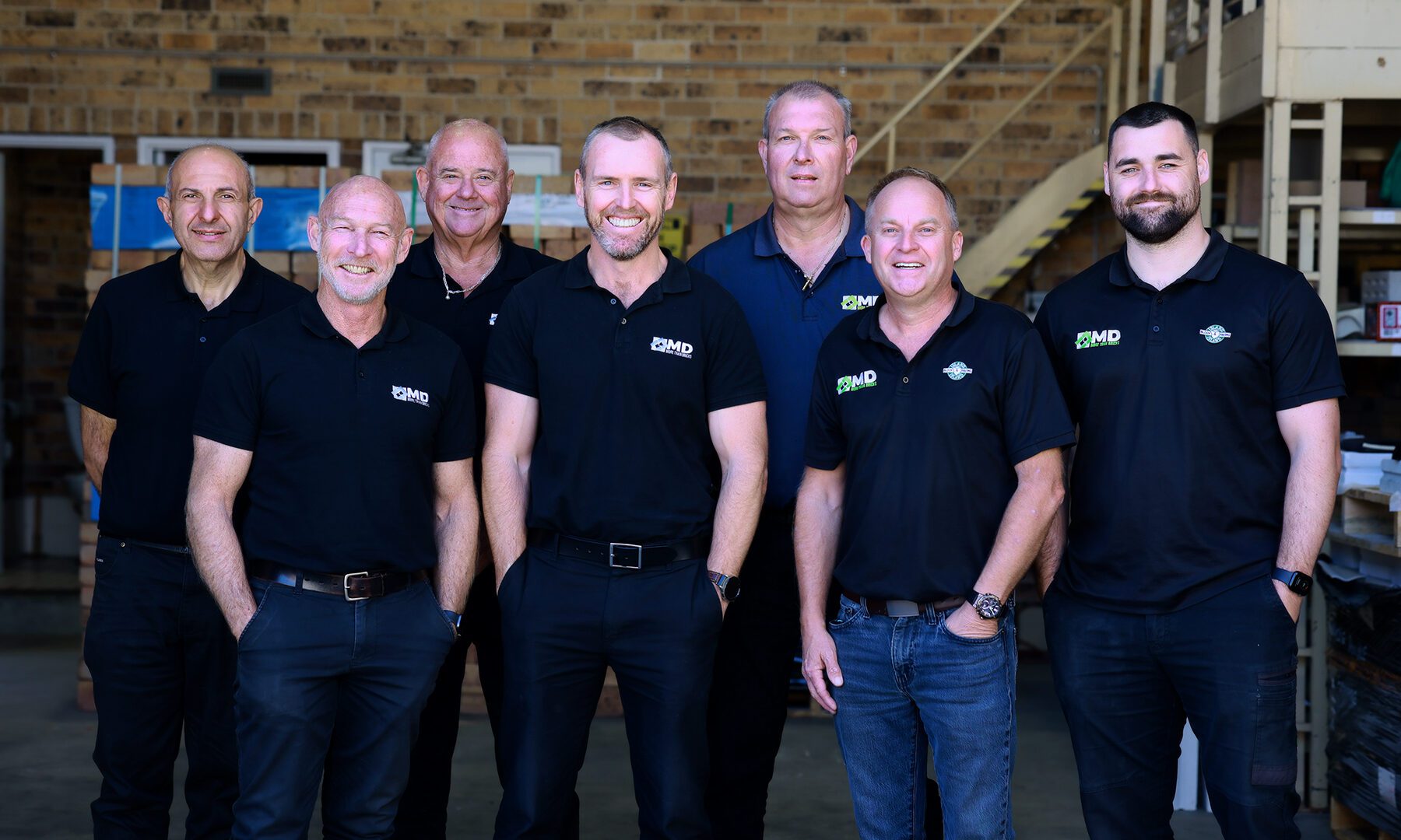 Seven men wearing black shirts stand smiling in a warehouse-like setting with brick walls and stairs in the background at MD Brick Central Coast.