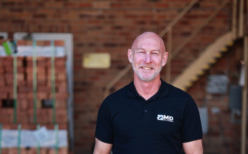 A smiling Martin Driene with a shaved head wears a black polo shirt with an MD logo, standing in a warehouse with stacked bricks in the background.